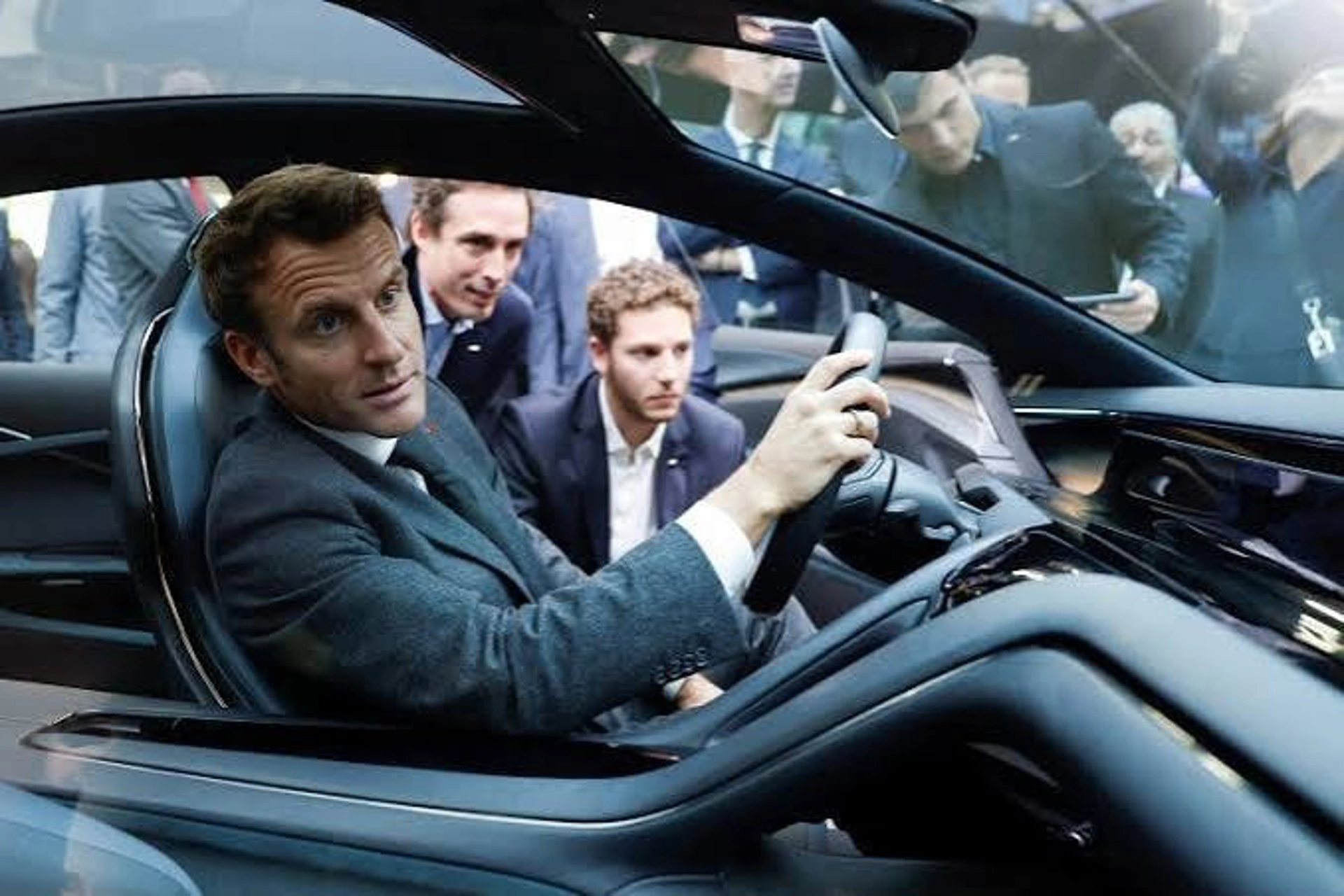 Emmanuel Macron sentado en el asiento del conductor de un automóvil moderno de exposición en el Salón del Automóvil de París, rodeado de asesores y asistentes.