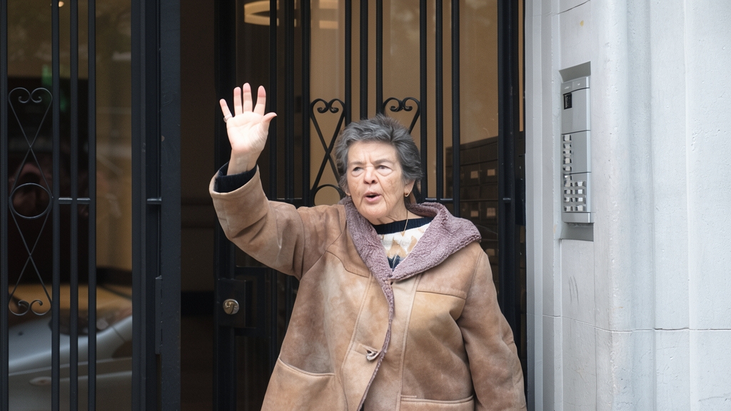 Fotografía de Maricarmen Abascal saludando con la mano levantada frente a la entrada de un edificio.