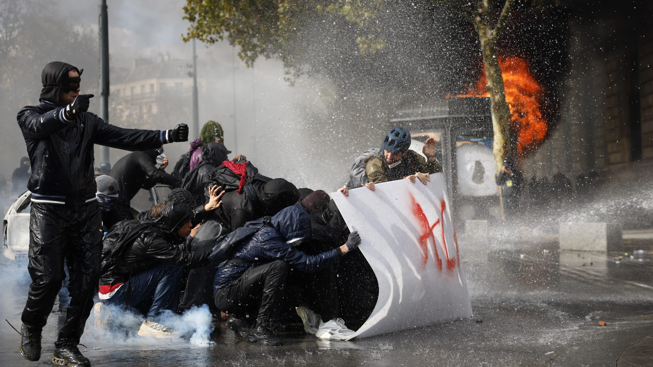 Manifestantes protegidos tras una pancarta soportando chorros de agua de cañones antidisturbios durante una protesta en París, con fuego y humo al fondo.