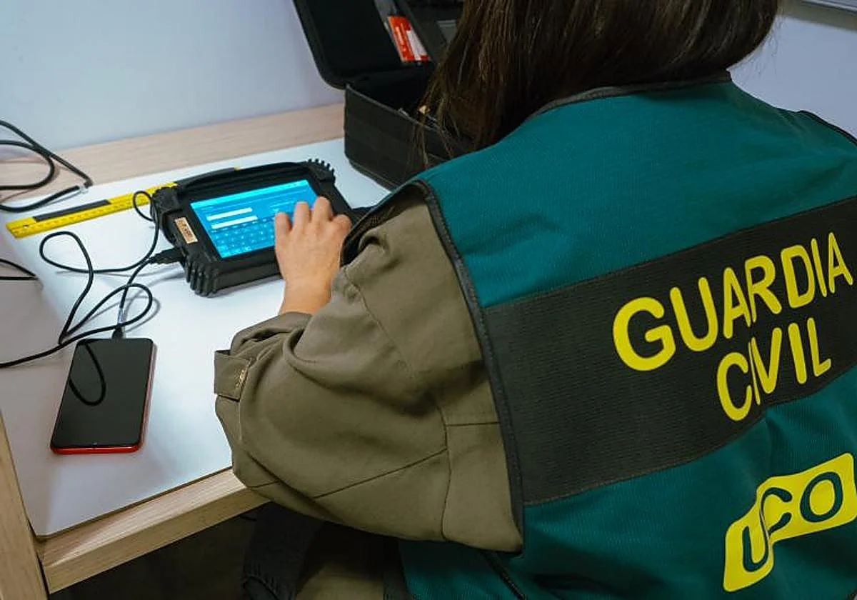 Agente de la Guardia Civil de la UCO investigando un ciberdelito analizando un dispositivo electrónico y un teléfono móvil en una mesa.