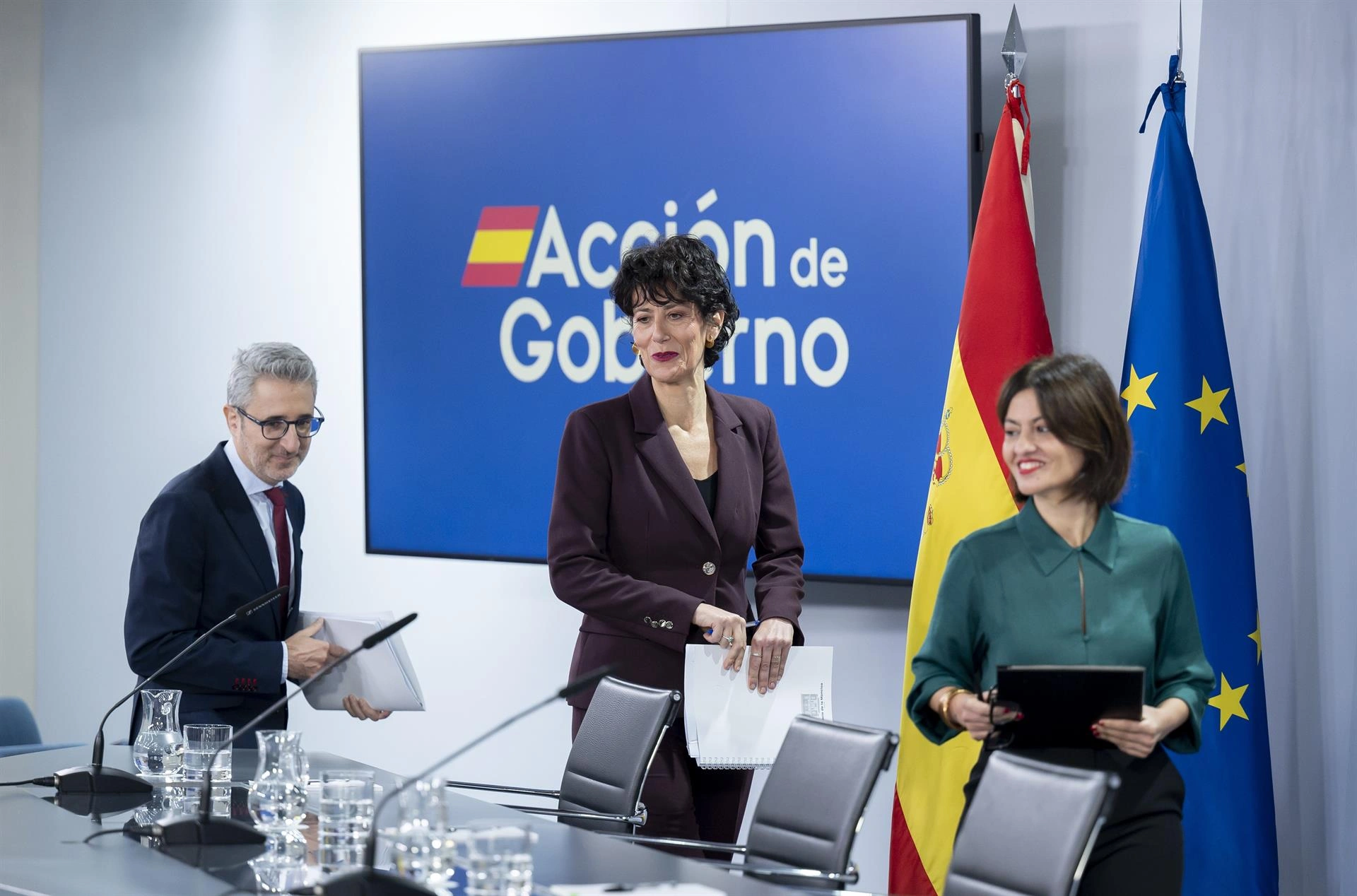 Composición de imágenes de una rueda de prensa con el logo de Acción de Gobierno, mostrando a un hombre con gafas, una mujer con traje oscuro y otra con camisa verde, junto a banderas de España y la UE.