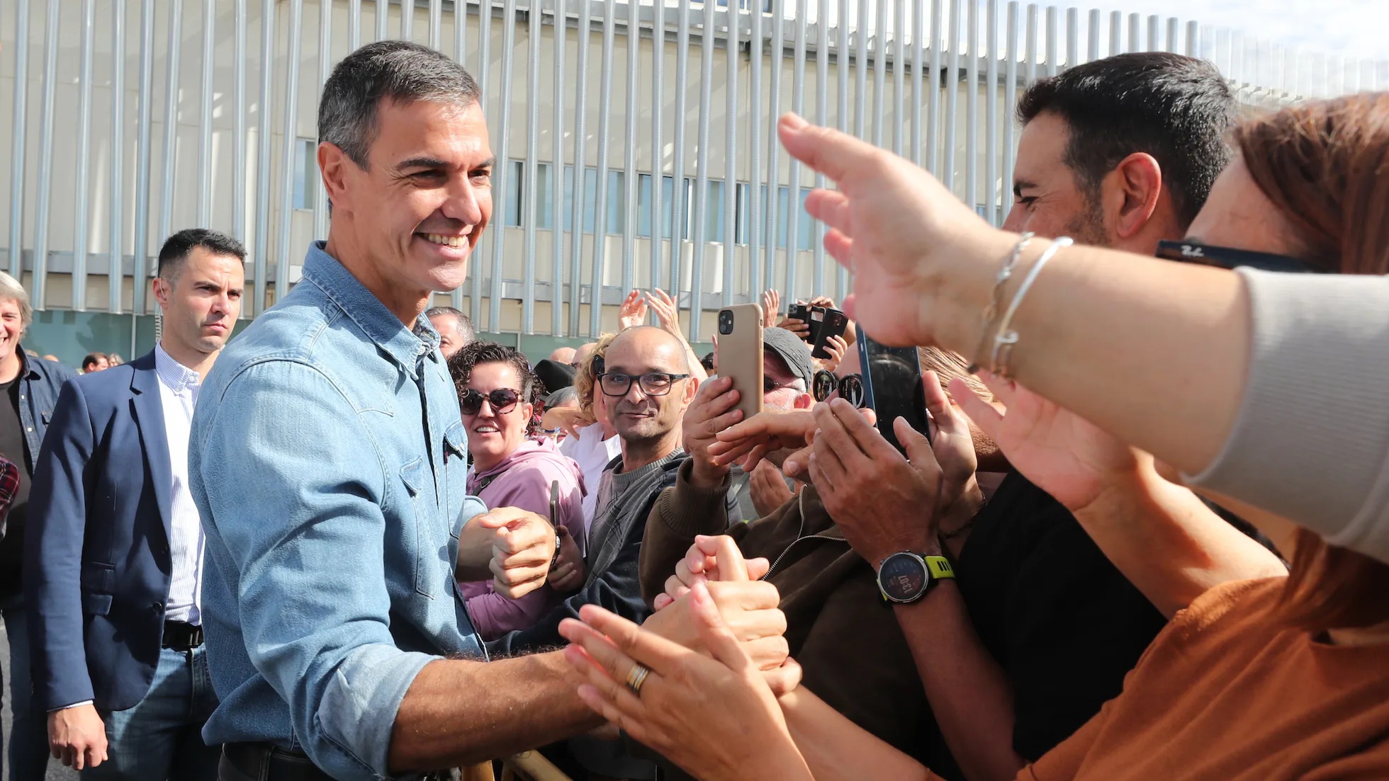 Pedro Sánchez saluda efusivamente a simpatizantes durante un acto público al aire libre, rodeado de una multitud que le toma fotografías con teléfonos móviles.