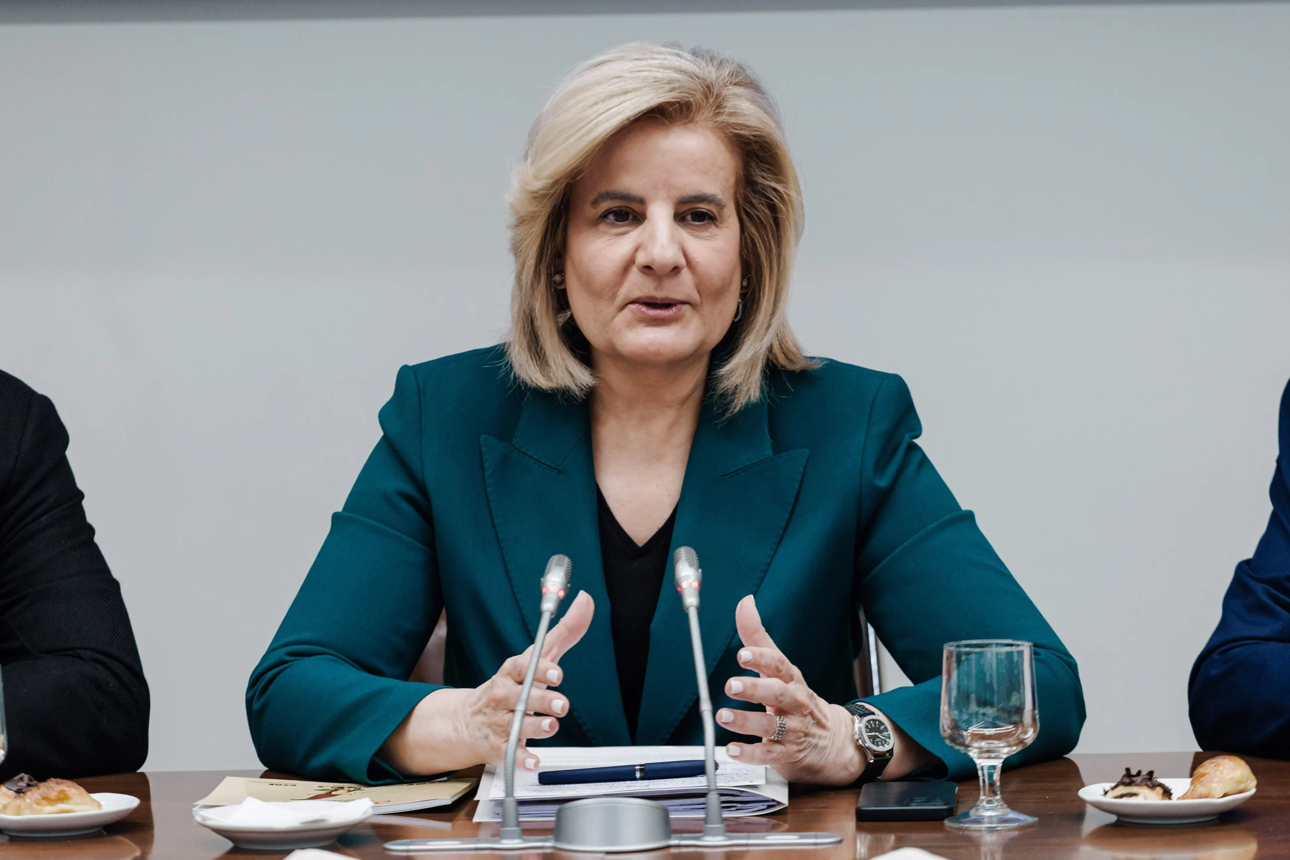 Fotografía de Fátima Báñez sentada en una mesa de conferencias con un traje verde oscuro y micrófonos delante.