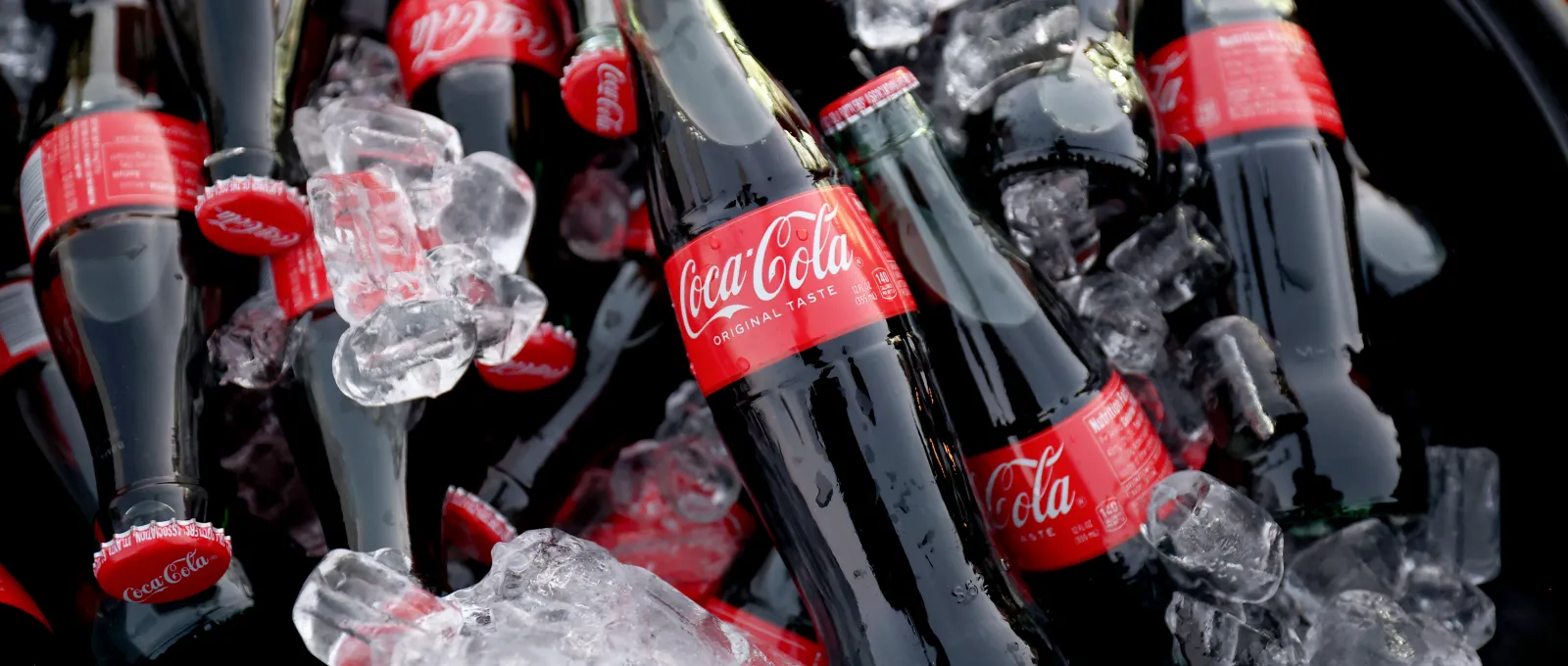 Varias botellas de Coca-Cola sumergidas en cubitos de hielo en una cubitera negra.