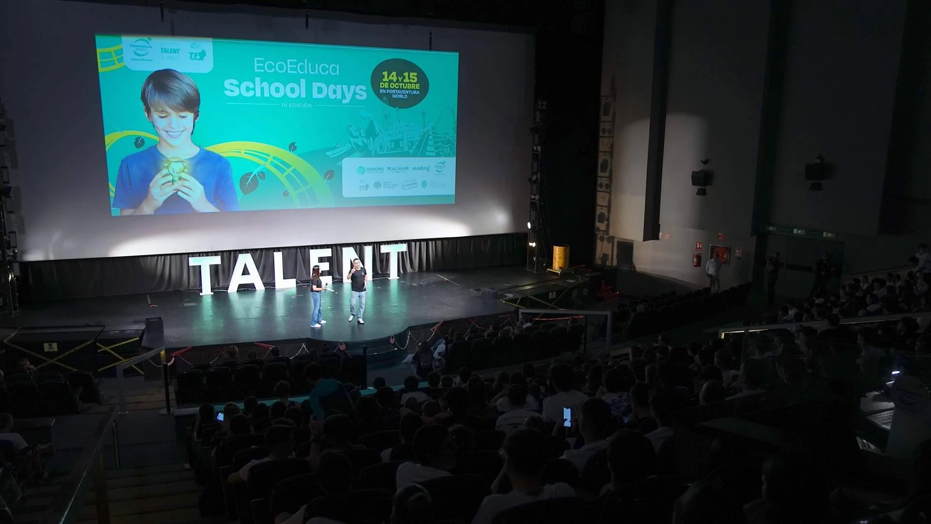 Fotografía editorial de la cuarta edición de PortAventura World Talent en un auditorio oscuro, con una gran pantalla de proyección que muestra el logotipo de EcoEduca School Days y dos ponentes en el escenario iluminado frente al público.