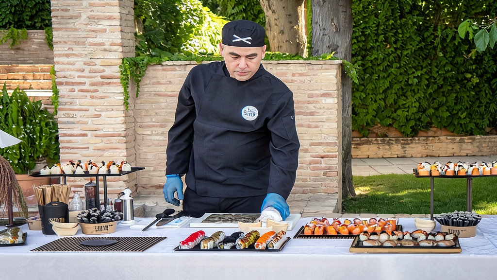 Chef de sushi preparando piezas en directo ante una mesa con bandejas de sushi variadas y decoración exterior.