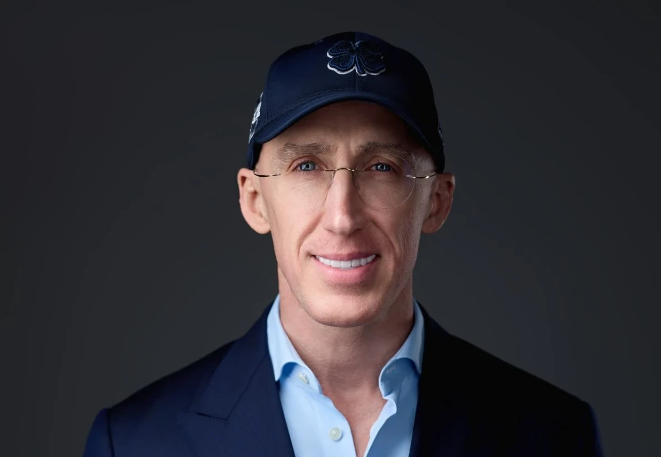 Retrato de Uri Poliavich con gorra azul con un trébol bordado, gafas minimalistas, camisa azul claro y chaqueta oscura frente a fondo gris.