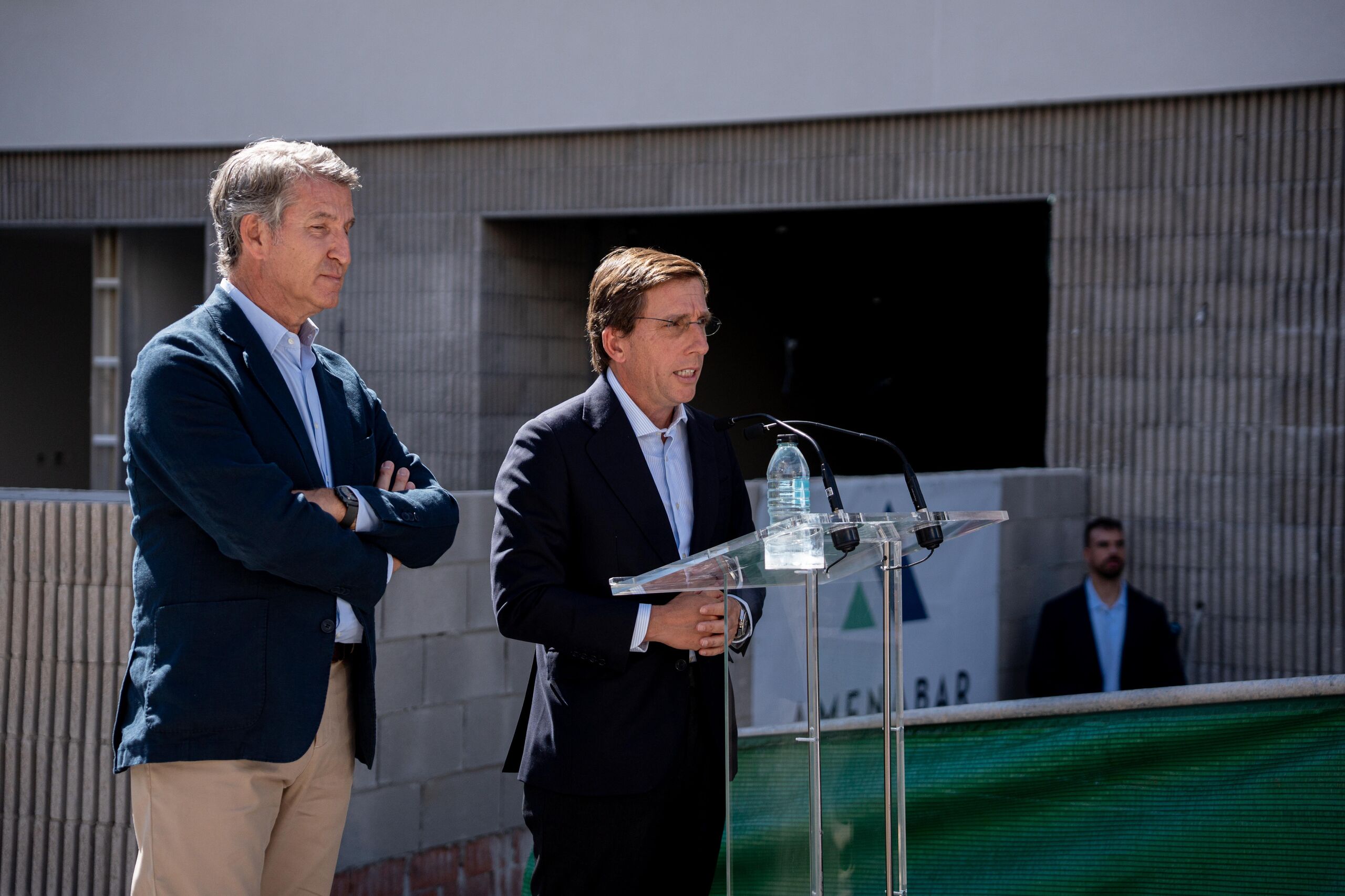 José Luis Martínez-Almeida hablando en un atril de cristal al aire libre, con Pedro Sánchez de pie a su lado con los brazos cruzados, frente a un edificio moderno de hormigón.